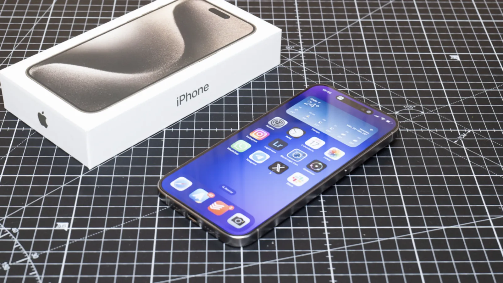 Fonctionnalités iOS 27 qui devraient devenir réalité sous Android 4 iPhone 15 Pro avec son écran allumé, posé à côté de son emballage d'origine sur un tapis de découpe quadrillé.