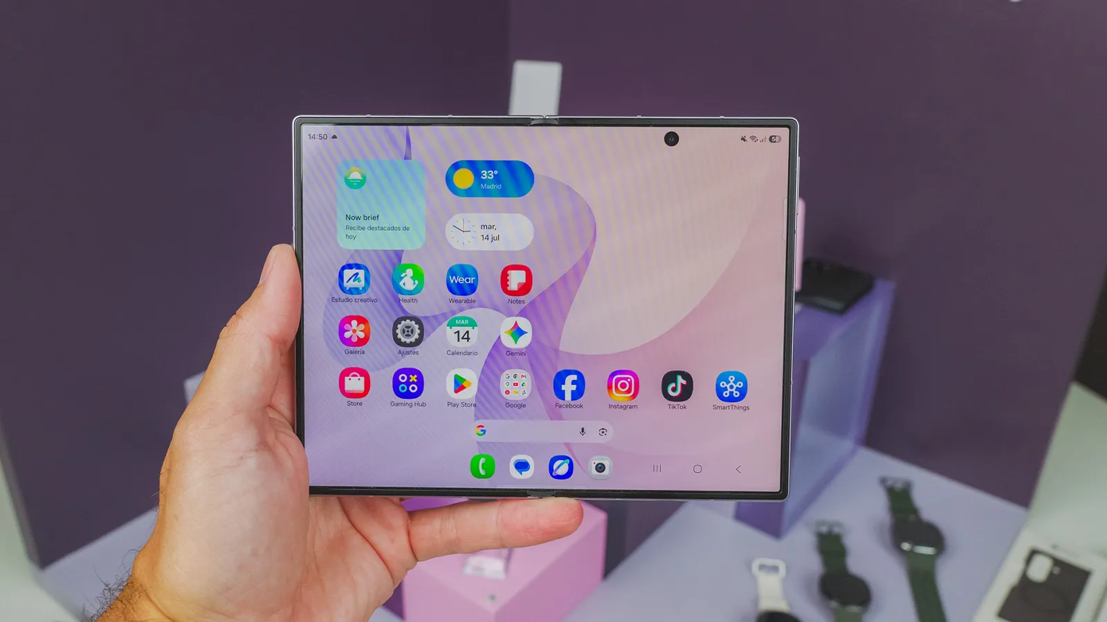 Main tenant un Samsung Galaxy Z Fold8 ouvert et montrant l'écran principal avec des icônes d'applications et des widgets.
